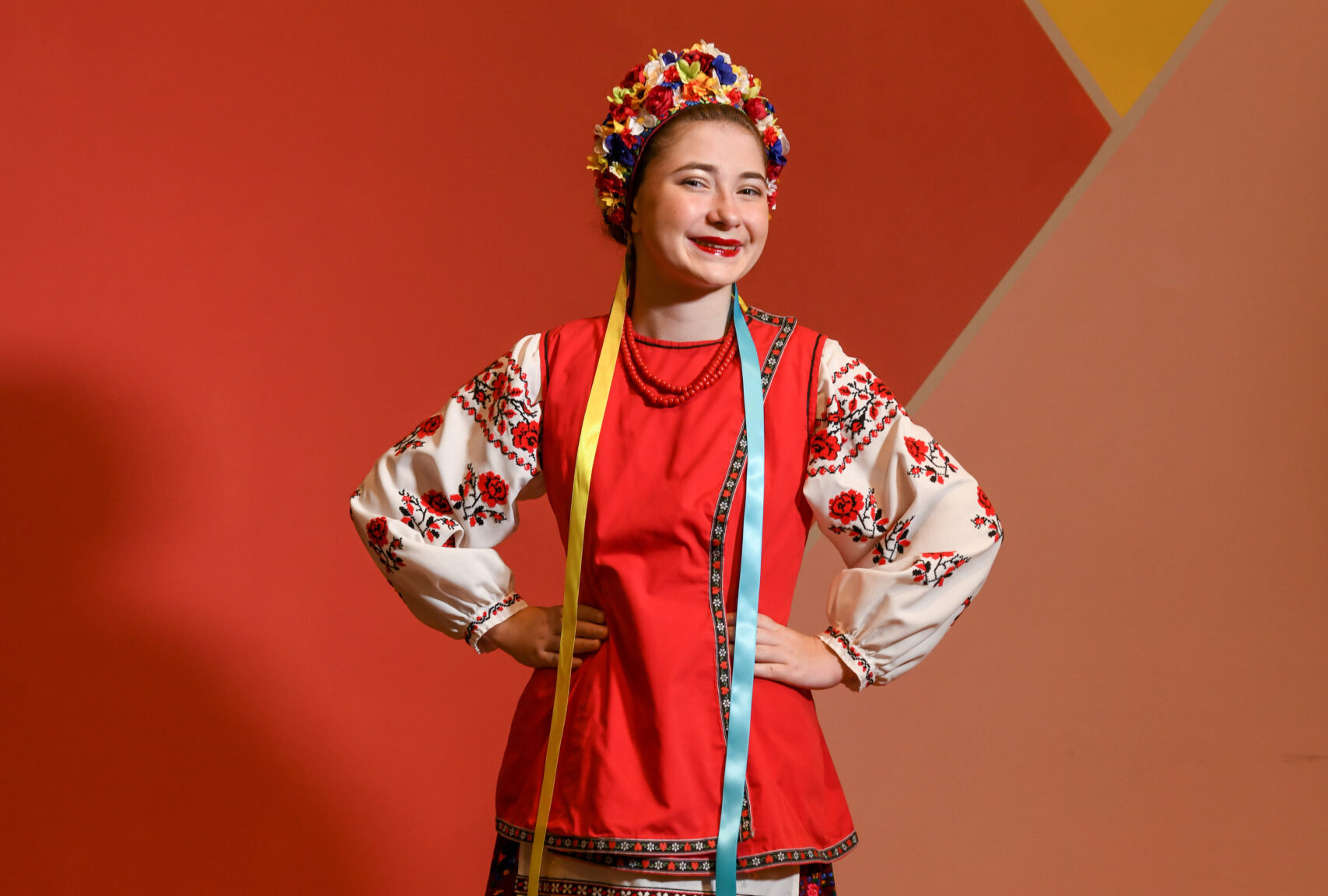 A young female dancer wears an embroidered shirt, a red apron, and a flower headpiece