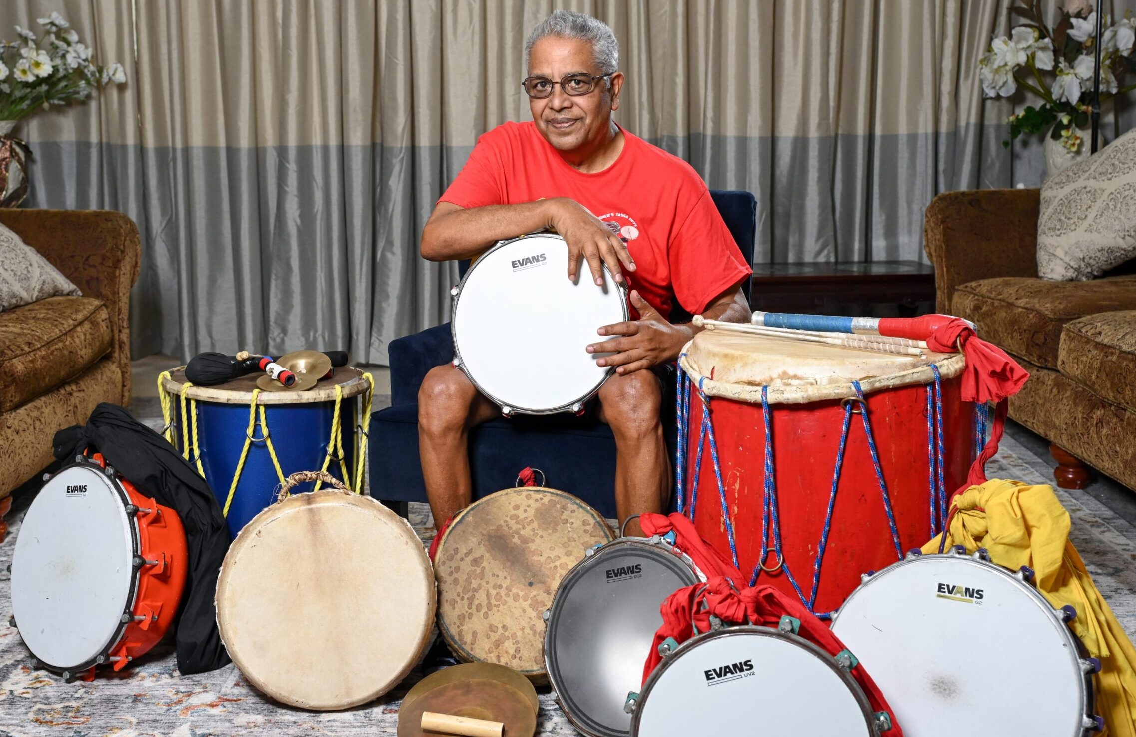 Romeo Ragbir sits surrounded by drums of various sizes. He holds a drum in his hands on his lap.