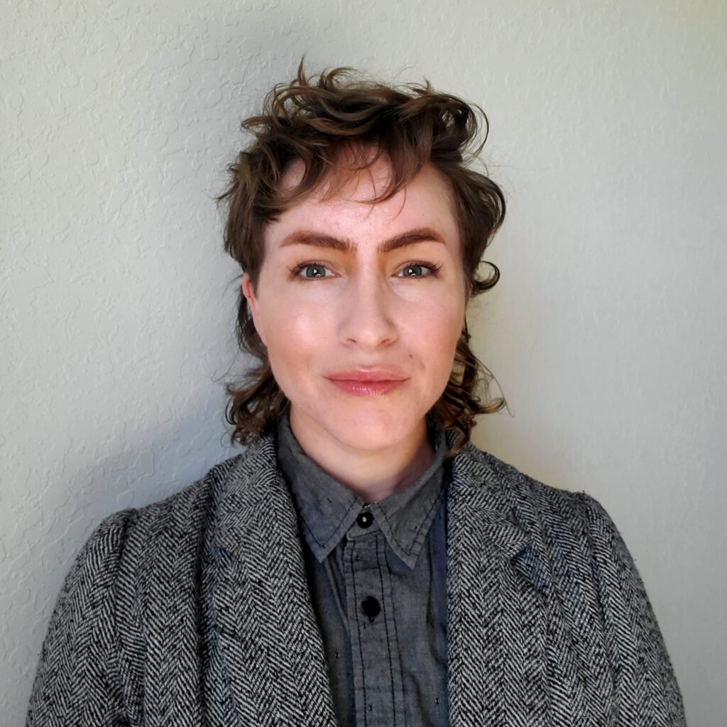A person with short brown curly hair stands in front of a grey background. They are wearing a dark grey suit jacket and grey button up shirt.