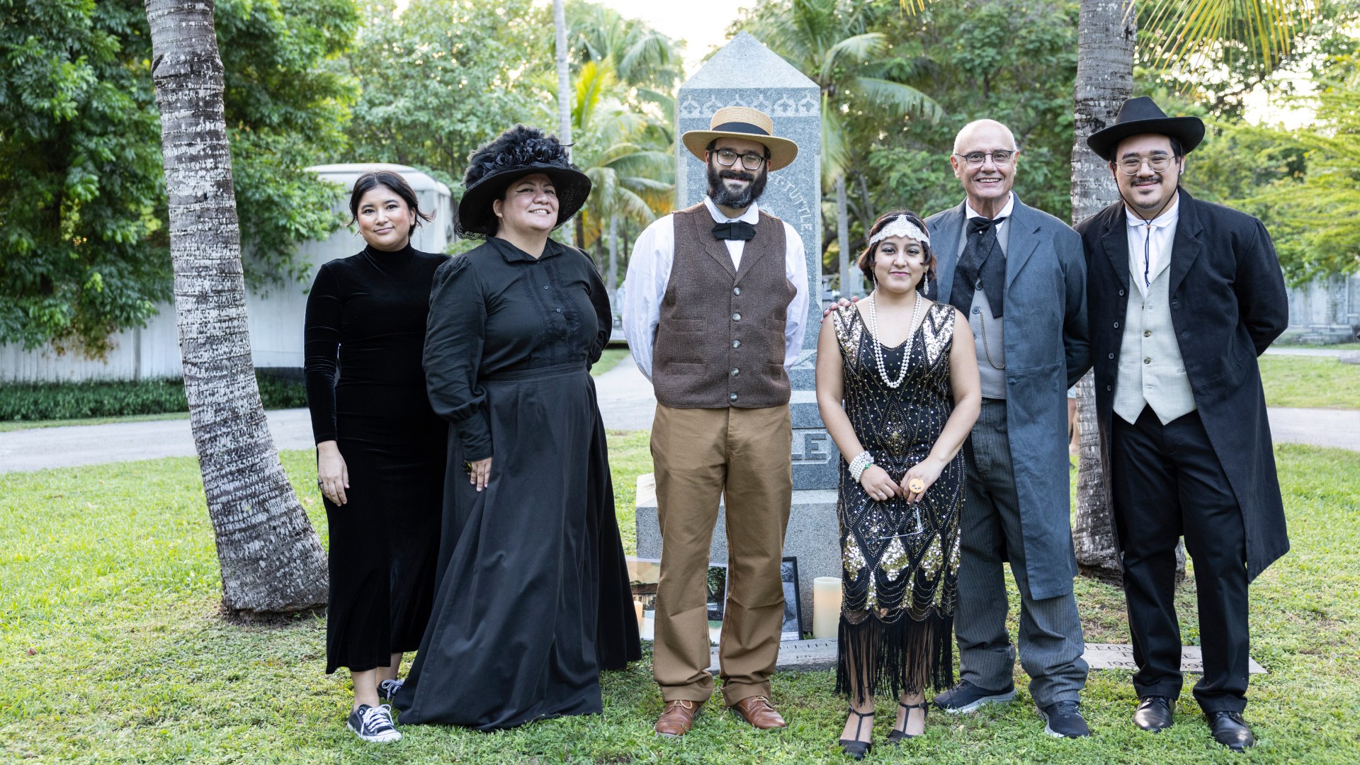 A group of people dressed in historical costumes stand together in front of a tall headstone inside of a cemetery. Green grass and trees can be seen in the background.