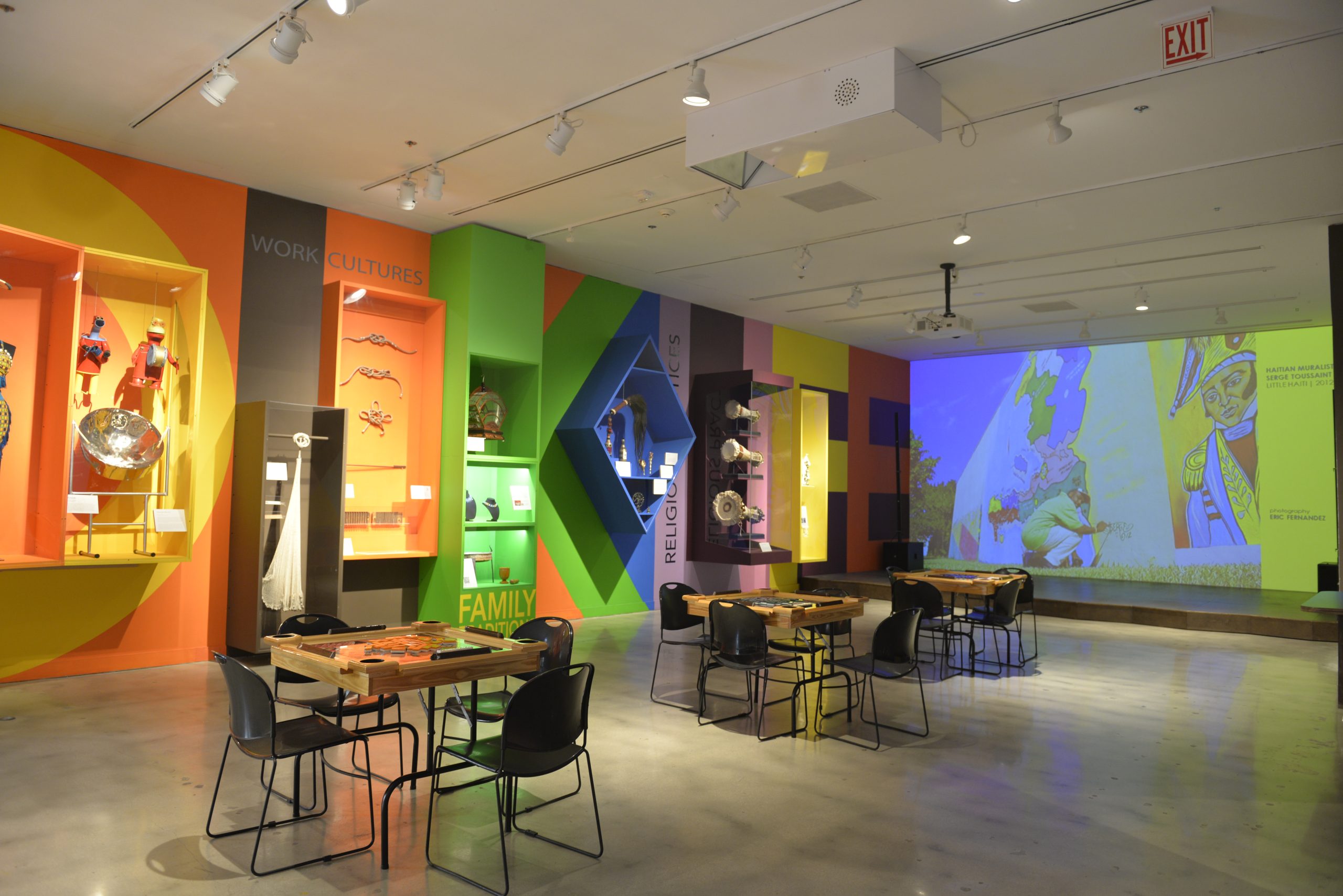 Domino tables sit in the middle of the room with objects encased along one wall. The other wall features a large projection with a photo of a man painting a mural.