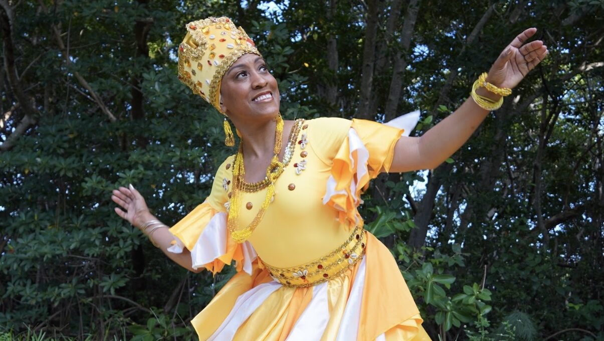 A woman poses with her arms lifted wearing a yellow crown and dress. She is standing in front of trees.