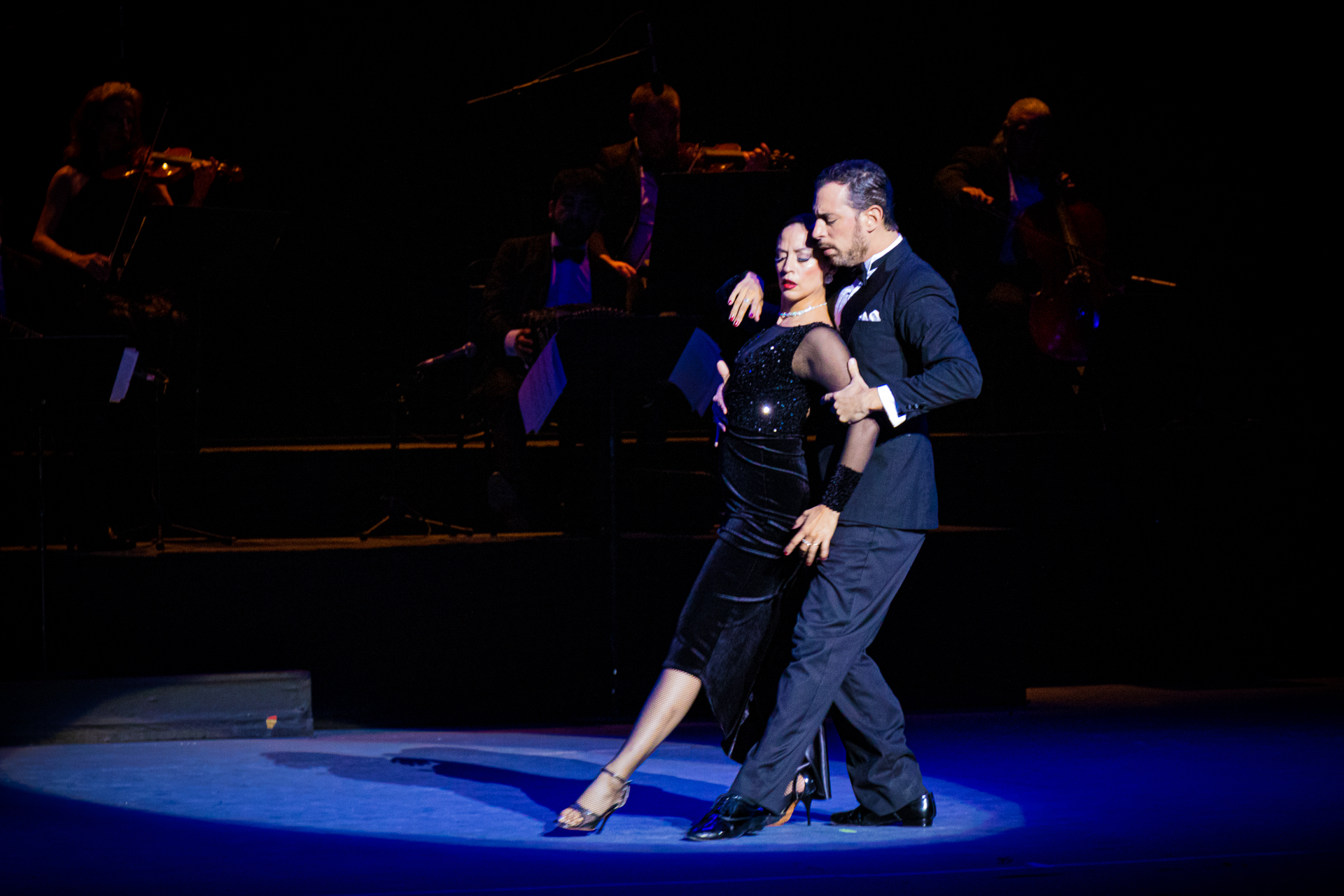 A man and woman are dancing tango. The woman stands with her back as he grabs her arm. They each have their left leg pointing forward.
