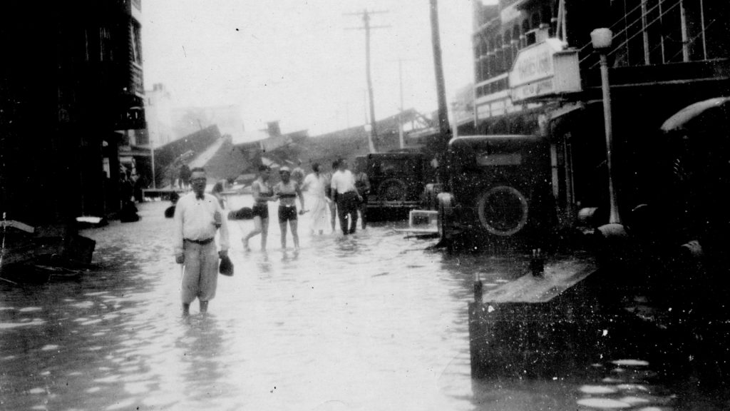 Ocean Drive after Hurricane, September 1926. Shows a street flooded by the 1926 Miami Hurricane. People stand in water that reaches above their ankles. A pair in the center are seen wearing swimwear. Debris is seen along the road and blocking the street in the distance. The entrance to Hardie’s Casino is seen to the right. Miami Beach, Fla. Miami News Collection. Museum of Miami, 1989-011-7760.